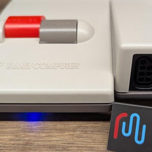 May include: A white Nintendo Family Computer (NES) console with a red power button and a gray reset button. The console has two controller ports on the right side. The console is sitting on a wooden surface.