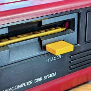 Puede incluir: Primer plano de un sistema de disco Nintendo Family Computer rojo y negro. Un disco amarillo está insertado en la ranura de la unidad, con un botón de expulsión amarillo. El texto "イジェクト" es visible.
