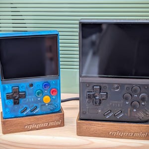 May include: Two handheld retro gaming consoles, one blue and one black, with a screen, buttons, and a directional pad. Both consoles are on wooden stands with the text "Miyoo Mini" printed on them.