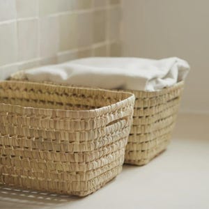 May include: Two woven rectangular baskets. One basket is in the foreground, and the other is behind it. The baskets are made of a light brown material. The basket in the background contains a white folded cloth. The baskets are on a white surface.