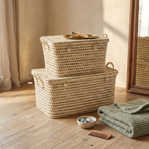May include: Two rectangular woven storage baskets, one stacked on top of the other. The baskets are made of a light-coloured natural material. A small bowl with stones, a wooden comb, and a folded green blanket are on the wooden floor.