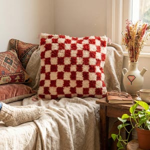 May include: A red and white checkered throw pillow is the focal point of this cozy scene. The textured pillow sits among other pillows on a neutral-toned blanket. A small wooden table holds a vase of dried flowers and a potted plant.