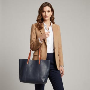 May include: A navy blue leather tote bag with tan handles. The bag is held by a person wearing a tan blazer, white shirt, and navy blue pants. The bag is a large, rectangular shape.