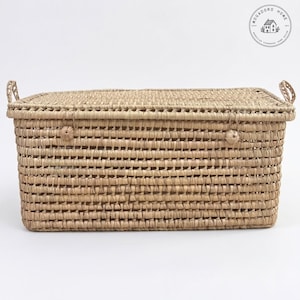 May include: A rectangular woven storage basket with a lid and handles. The basket is made from natural-coloured woven material. The lid features a woven pattern and is secured with a small woven knot. The basket is rectangular in shape and suitable for storage.