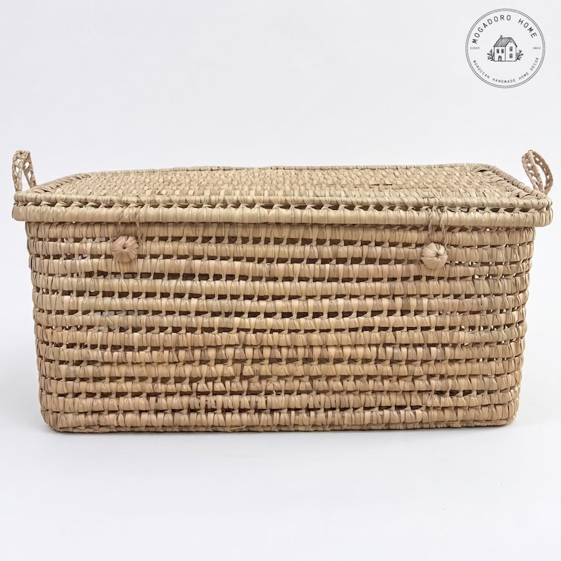 May include: A rectangular woven storage basket with a lid and handles. The basket is made from natural-coloured woven material. The lid features a woven pattern and is secured with a small woven knot. The basket is rectangular in shape and suitable for storage.