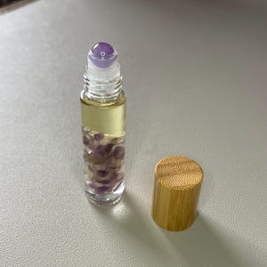 May include: A clear glass roller bottle with a purple gemstone roller ball and a bamboo lid. The bottle is filled with a clear liquid and purple gemstones.