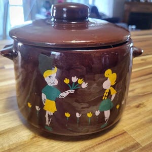 May include: A brown ceramic lidded casserole dish with handles. The dish features a painted design of two figures, one offering flowers to the other. Yellow, white, and green accents decorate the dish. The lid has a small knob.