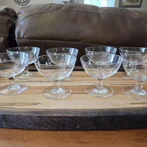May include: A set of seven clear glass coupes with short stems and circular bases, each featuring a delicate etched starburst pattern on the bowl. The glasses are arranged on a wooden serving tray with a light-colored center and dark edges.