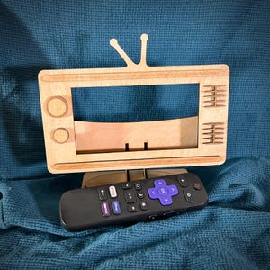 May include: A wooden TV stand with a cutout for a phone or tablet. The stand is painted brown and has a retro design. A black remote control with buttons for Netflix, Hulu, Disney+, and Amazon Prime is sitting in front of the stand.