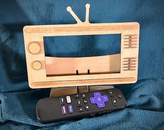 May include: A wooden TV stand with a cutout for a phone or tablet. The stand is painted brown and has a retro design. A black remote control with buttons for Netflix, Hulu, Disney+, and Amazon Prime is sitting in front of the stand.