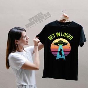Puede incluir: Camiseta negra con un gráfico de una vaca siendo abducida por un OVNI, sobre un fondo de puesta de sol. El texto "GET IN LOSER" está impreso en verde neón sobre el gráfico.