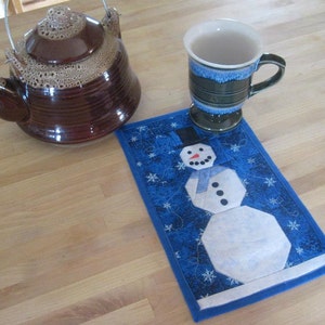 Pode incluir: Um descanso de copo acolchoado azul e branco com tema de boneco de neve com um bule marrom e uma caneca verde em uma mesa de madeira.