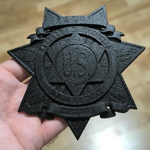 May include: A black, star-shaped badge with a textured finish. The center features a circular emblem with the letters US. A banner at the top displays the word ZAXON, and a banner at the bottom contains additional text. The badge has a layered, relief design with intricate details throughout the surface.
