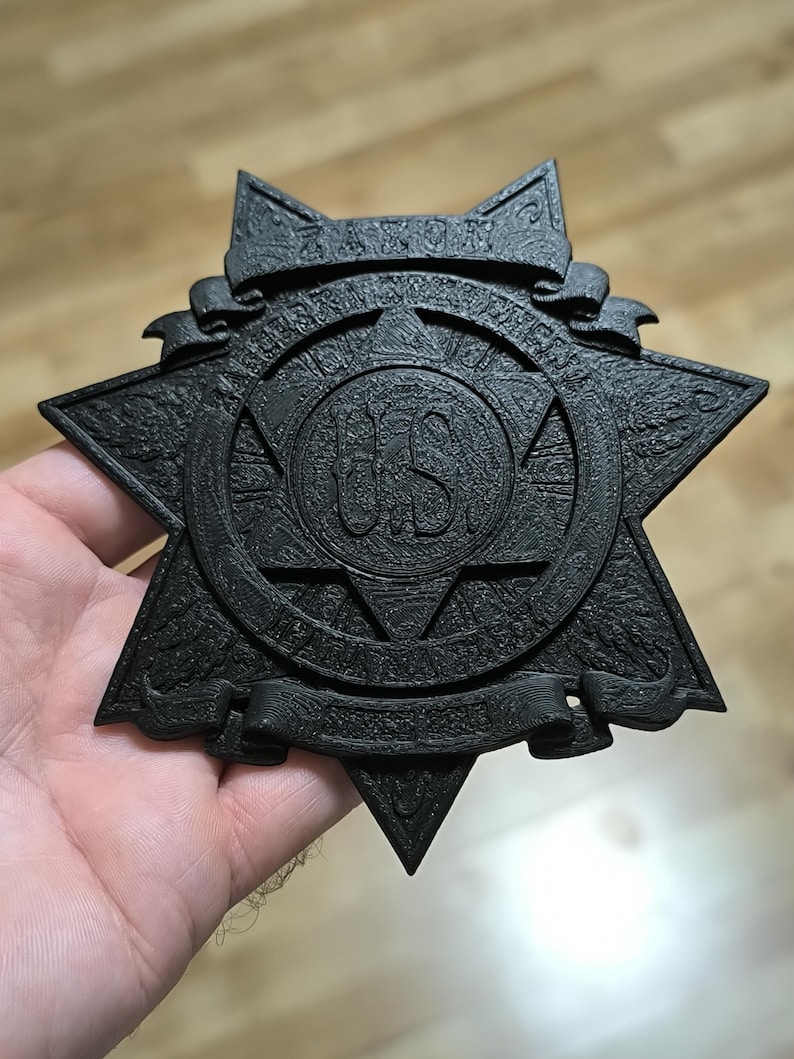 May include: A black, star-shaped badge with a textured finish. The center features a circular emblem with the letters US. A banner at the top displays the word ZAXON, and a banner at the bottom contains additional text. The badge has a layered, relief design with intricate details throughout the surface.