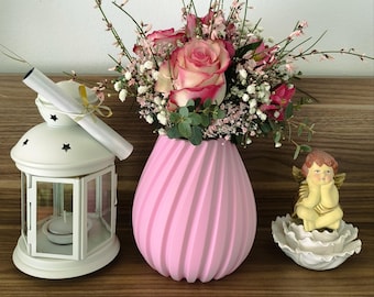 May include: A pink ceramic vase with a swirl design holds a bouquet of pink roses and white baby's breath. The vase is on a wooden surface next to a white lantern and a small white angel figurine.