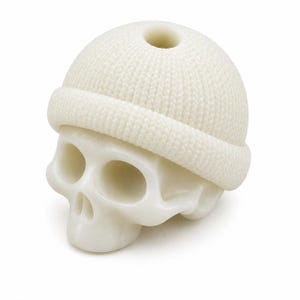 May include: A white, smooth-surfaced skull sculpture wearing a textured, knit-patterned beanie cap. The beanie features a prominent folded cuff and a small circular opening at the very top. The skull has hollow eye sockets and a nasal cavity.