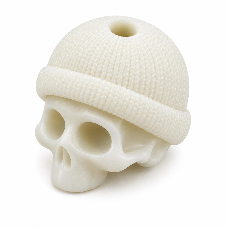 May include: A white, smooth-surfaced skull sculpture wearing a textured, knit-patterned beanie cap. The beanie features a prominent folded cuff and a small circular opening at the very top. The skull has hollow eye sockets and a nasal cavity.