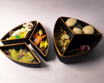 May include: A set of three black serving bowls with gold-toned rims, featuring a triangular, segmented design. The bowls are filled with various snacks, including assorted gummy candies, small crackers, and individually wrapped treats.