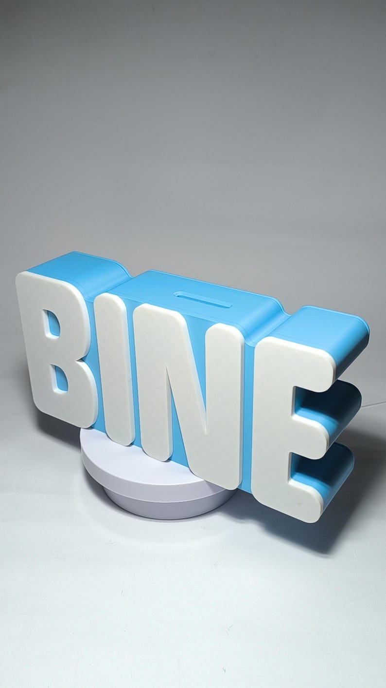 May include: A blue and white piggy bank in the shape of the word "BINE". The letters are white with a blue outline and a slot for inserting money. The bank sits on a white circular base.