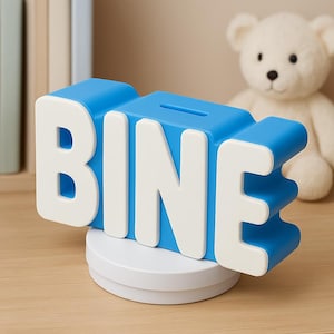 May include: A blue and white piggy bank in the shape of the word "BINE". The letters are white with a blue outline. The piggy bank sits on a white circular base. A slot is visible on the top. A white teddy bear is in the background.