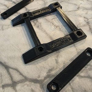 Peut inclure: Composants noirs imprimés en 3D pour Bash Rack. L'image montre un cadre carré avec le texte "Bash Rack" et deux petites pièces rectangulaires. Toutes les pièces ont des trous pour l'assemblage. Les articles sont sur une surface texturée.