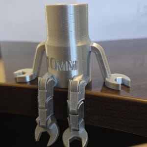 May include: A silver 3D printed robot figurine with articulated legs and arms. The robot is sitting on a wooden surface. The robot's body is a cylinder with the text "10MM" printed on it. The robot's arms end in wrench shapes.