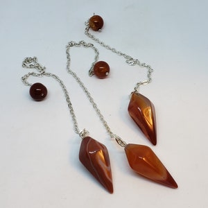 Può includere: Tre pendenti in cristallo di corniola con catene d'argento e perle di corniola. I pendenti sono sfaccettati e hanno un colore marrone rossiccio.