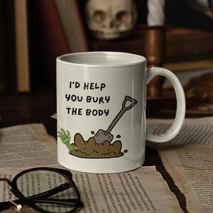 May include: White ceramic coffee mug with a black and white illustration of a hand digging a hole with a shovel. The text "I'd help you bury the body" is printed on the mug.