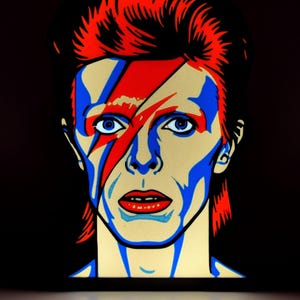 Può includere: Ritratto pop art di una persona con capelli rossi e un fulmine rosso e blu sul viso. Il viso è dipinto con accenti blu e rossi e lo sfondo è nero.