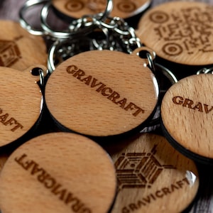 Könnte beinhalten: Eine Sammlung von Holz-Schlüsselanhängern mit silbernen Schlüsselringen. Die Schlüsselanhänger sind rund und mit dem Wort "GRAVICRAFT" graviert. Einige Schlüsselanhänger haben ein geometrisches Design oder einen QR-Code. Das Holz hat eine hellbraune Farbe.
