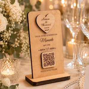 Könnte beinhalten: Ein Holzschild für eine Hochzeit mit einem herzförmigen Akzent, graviert mit Namen und Datum. Das Schild enthält einen QR-Code und die Worte "TEILE EURE SCHÖNSTEN MOMENTE MIT UNS" und "FOTOS HIER TEILEN". Das Schild steht auf einem Tisch mit Blumen und Kerzen.