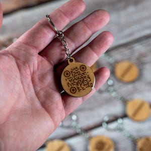 Könnte beinhalten: Ein Holzschlüsselanhänger mit einem kreisförmigen Anhänger mit einem QR-Code und zwei konzentrischen Kreisen. Der Schlüsselanhänger ist an einer silbernen Kette und einem Verschluss befestigt. Der Anhänger wird in einer Hand gehalten.
