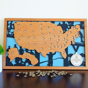 May include: Wooden United States map with state-shaped pieces, framed in a light brown wood. The background features a blue sky and black silhouettes of runners. A medal with the text "Marathon Adventure Map" is attached. Small wooden pieces are scattered below.