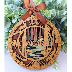 May include: A wooden ornament with a brown and natural wood finish. It features a detailed design of a boardwalk trail, a river, and a forest. The text "CONGAREE" is prominently displayed in the center. The ornament also includes the text "FIREFLIES BIOBLITZ 33.8 N 80.7 W ANCIENT FOREST CONGAREE RIVER BOARDWALK TRAIL".