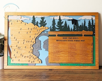 May include: A wooden map of Minnesota state parks with a sign that reads "Dave and Deb's Minnesota State Parks Map". The map is framed in wood and features a blue sky with white clouds, green trees, and a river. The map is decorated with small wooden circles representing each state park.