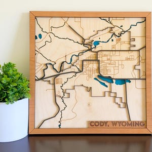 May include: A wooden map of Cody, Wyoming, framed in light brown wood. The map features detailed laser-cut streets, rivers, and lakes in shades of brown, black, and blue. The text "CODY, WYOMING" is carved at the bottom.