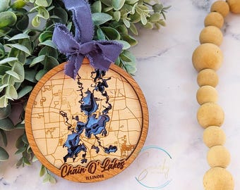 May include: Wooden ornament shaped like the Chain O' Lakes in Illinois. The ornament is light brown with a dark brown outline of the lakes. The text "Chain O' Lakes" and "Illinois" is engraved on the ornament.