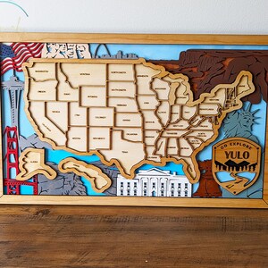 May include: A wooden wall art piece depicting a map of the United States with various landmarks, including the Golden Gate Bridge, the Statue of Liberty, and the White House. The map is surrounded by a wooden frame and features a blue and white background with the text "GO EXPLORE YULO" on a brown patch.