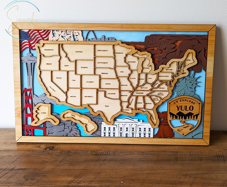 May include: A wooden wall art piece depicting a map of the United States with various landmarks, including the Golden Gate Bridge, the Statue of Liberty, and the White House. The map is surrounded by a wooden frame and features a blue and white background with the text "GO EXPLORE YULO" on a brown patch.