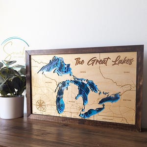 May include: Wooden framed map of the Great Lakes with blue layered water detail. The map includes state names and a compass rose. The frame is dark brown, and the text "The Great Lakes" is engraved.