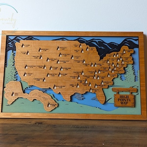 May include: A wooden map of the United States with the highest point in each state marked with a small wooden mountain peak. The map is framed in a light brown wood frame and has a blue and green background. The sign at the bottom reads "HIGH POINTS MAP".