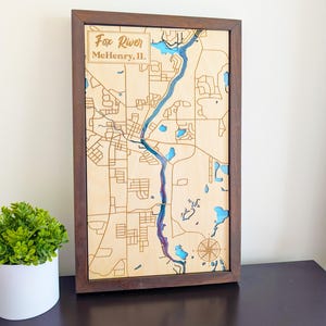 May include: Wooden framed map of Fox River, McHenry, IL, with laser-cut details. The river and lakes are highlighted in blue and purple. A compass rose is in the lower right corner. The frame is dark brown.