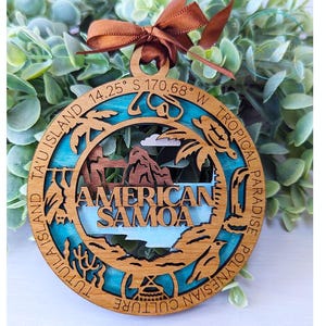 May include: Wooden ornament with a blue and green background. The ornament features a map of American Samoa with the text "American Samoa" and "Tutuila Island Ta'u Island 14.25° S 170.68° W Tropical Paradise Polynesian Culture".