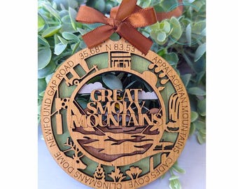 May include: A wooden ornament shaped like a circle with a green and brown design. The ornament features the text "Great Smoky Mountains" and the names of various locations in the Great Smoky Mountains National Park, including Clingmans Dome, Newfound Gap Road, and Cades Cove. The ornament is decorated with a brown ribbon.