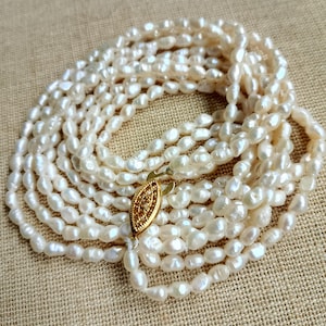 May include: A long, multi-strand necklace composed of creamy white, irregularly shaped pearls. The necklace is fastened with a gold-colored clasp. The pearls are arranged in multiple strands, creating a layered effect. The background is a neutral, textured surface.