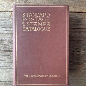 May include: A maroon-colored book titled "Standard Postage Stamp Catalogue" with gold lettering. The book's cover also includes the text "The Encyclopedia of Philately". The book is resting on a weathered wooden surface.