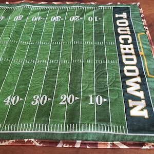 May include: Green football field quilt with white yard lines and numbers. The word "TOUCHDOWN" is printed vertically in white and gold. The quilt has a brown and tan football-themed border.