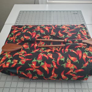 May include: A black fabric hot pad with a red chili pepper pattern. The hot pad is folded in half and has a wooden spoon inside.
