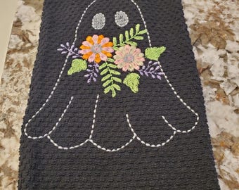 May include: A dark gray hand towel with an embroidered ghost design. The ghost has embroidered eyes and a floral bouquet in shades of orange, pink, and green. The towel is made of a textured fabric.