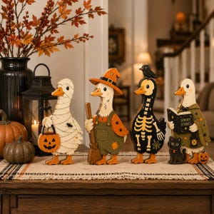 May include: Four wooden duck figurines decorated for Halloween. One is wrapped like a mummy holding a pumpkin bucket, one wears a witch hat and holds a broom, one features a skeleton print with a bird on its head, and one wears a cape reading Spooky Stories.